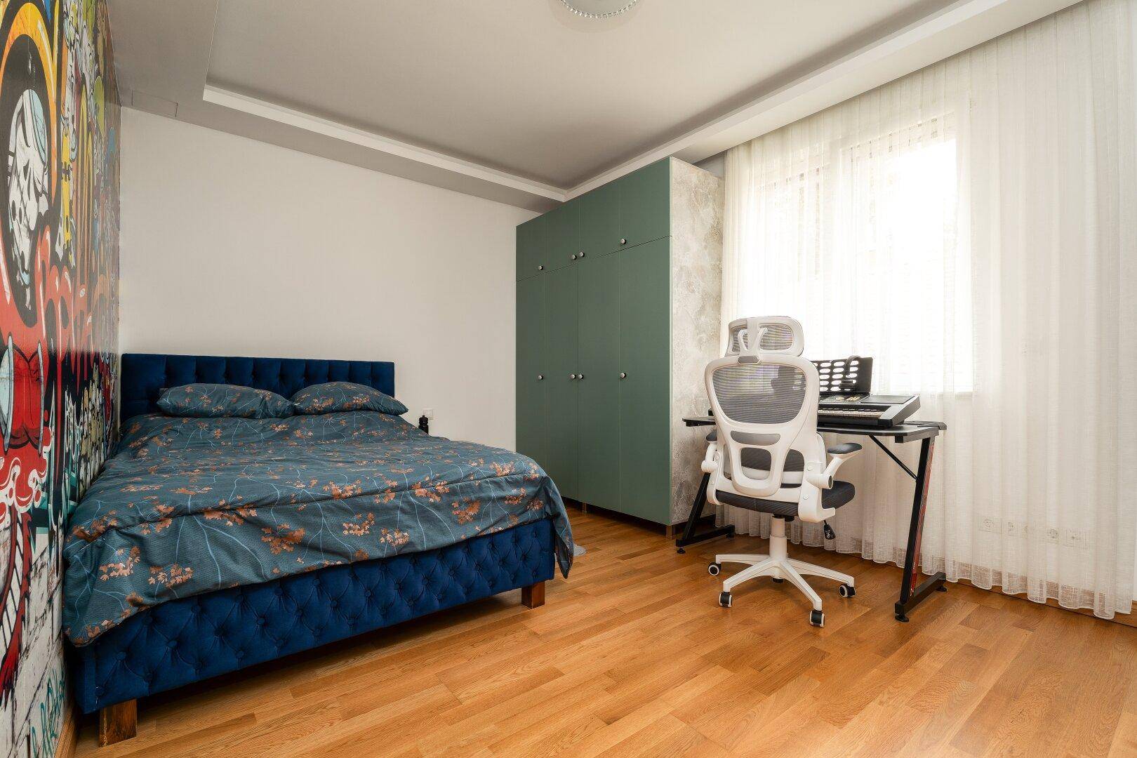 Jugendliches Schlafzimmer mit blauem Bett, grünem Kleiderschrank und farbenfroher Graffiti-Wand.