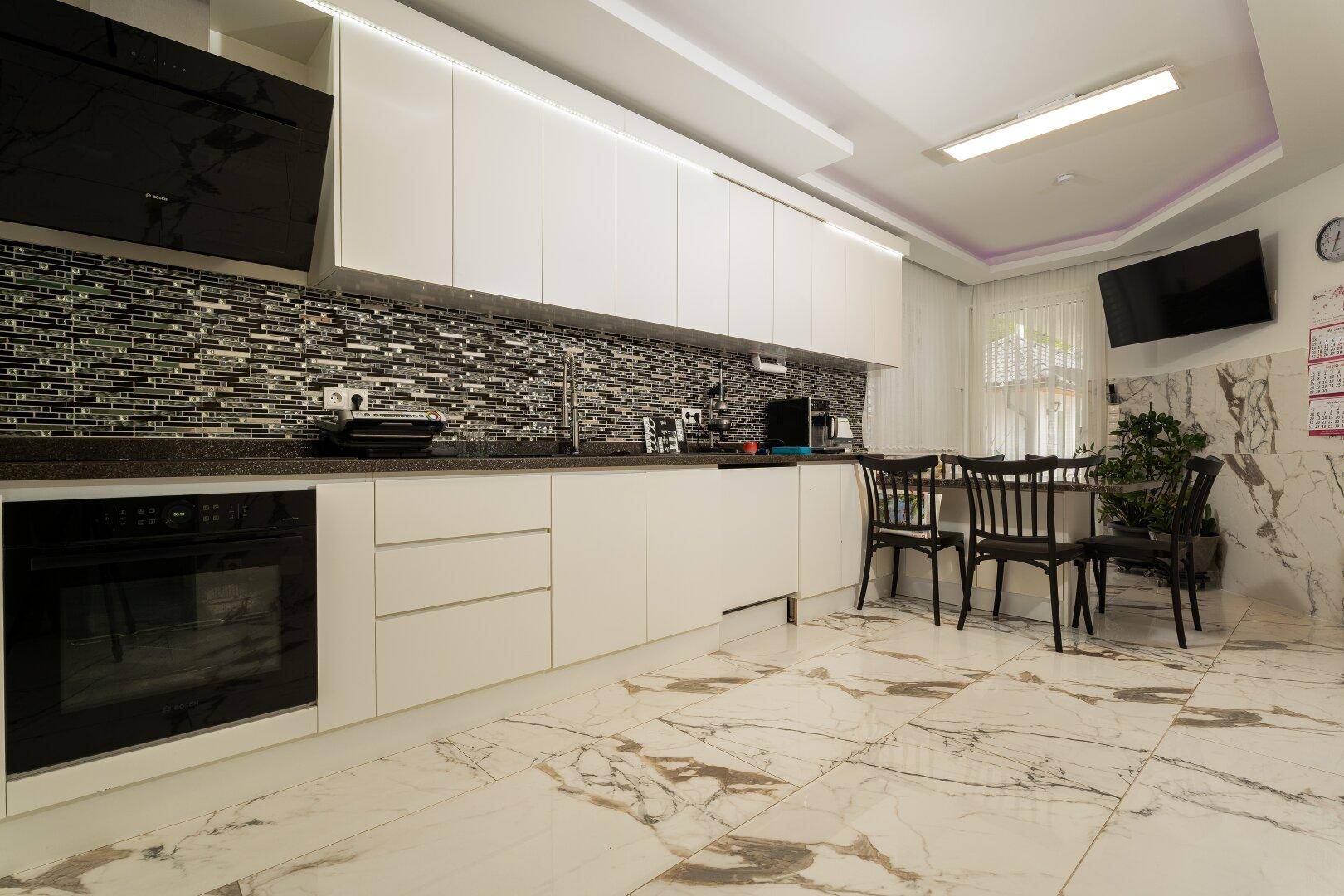 Moderne Küche mit weißen Schränken, schwarzem Mosaik-Spritzschutz und einem Essbereich mit dunklen Stühlen.