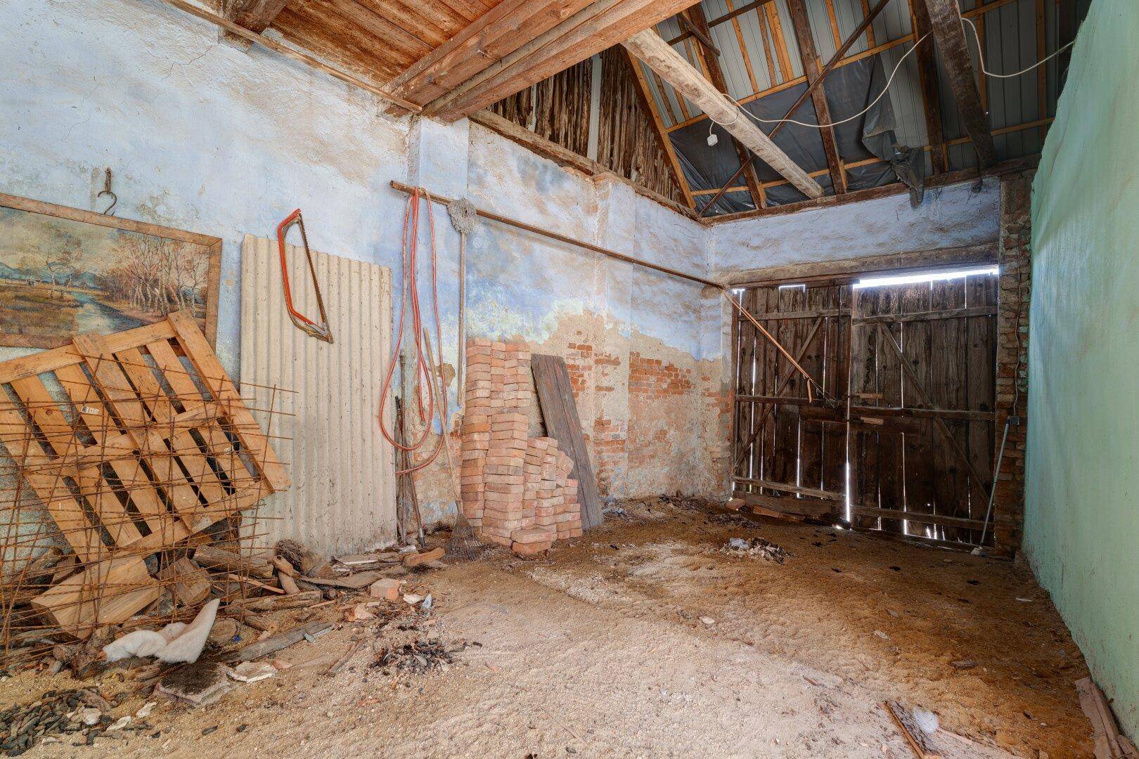 Rustikaler Scheunenraum mit Holzbalken, Ziegelwänden und Erdboden.