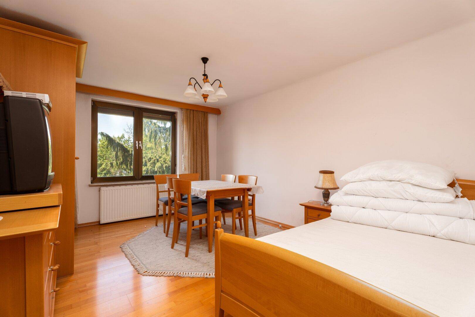 Helles Schlafzimmer mit Holzboden, einem Bett und einem kleinen Essbereich mit Blick ins Grüne.