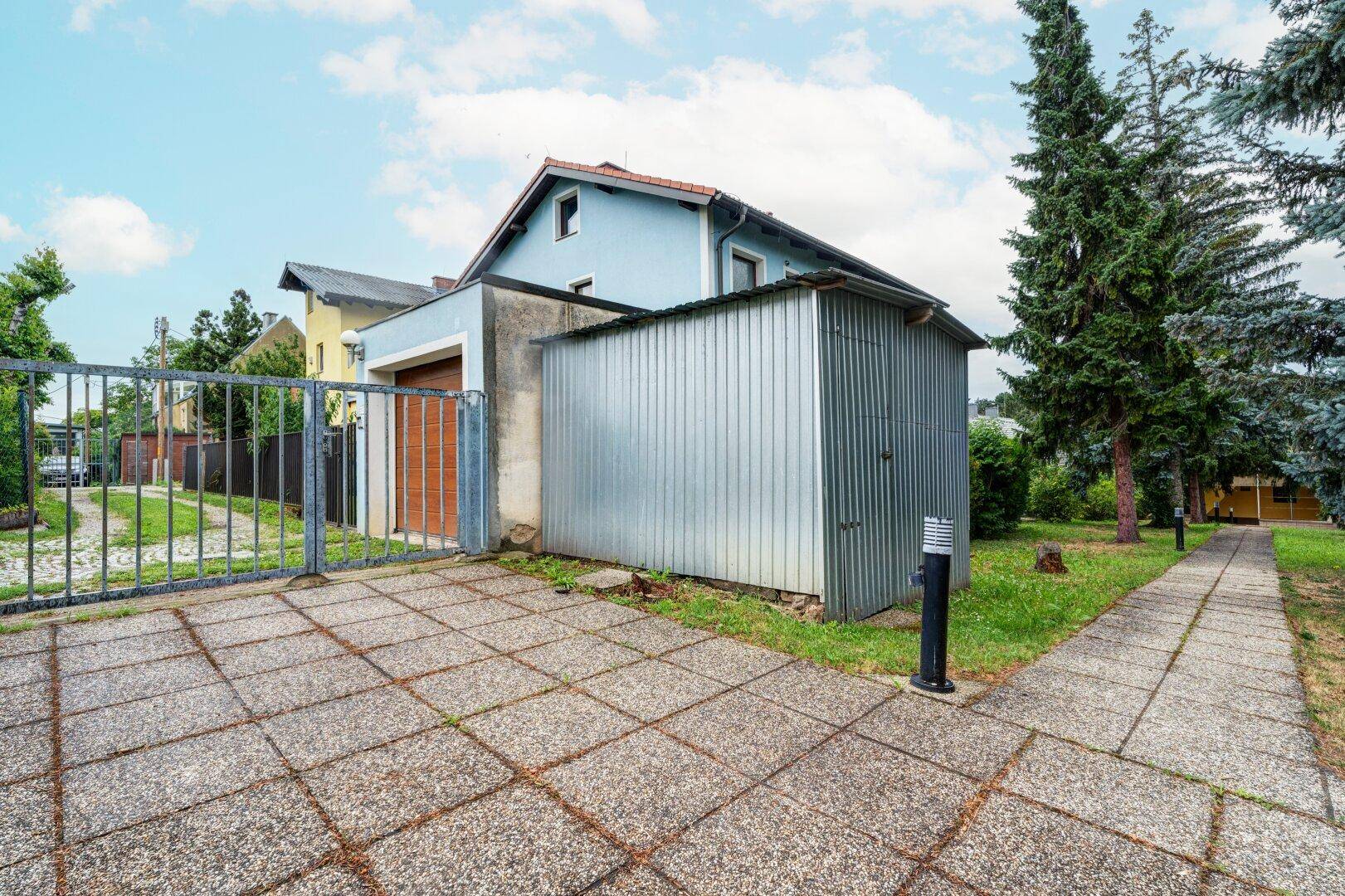 Einfahrt mit gepflastertem Bereich, Metallschuppen und einem blauen Haus im Hintergrund.