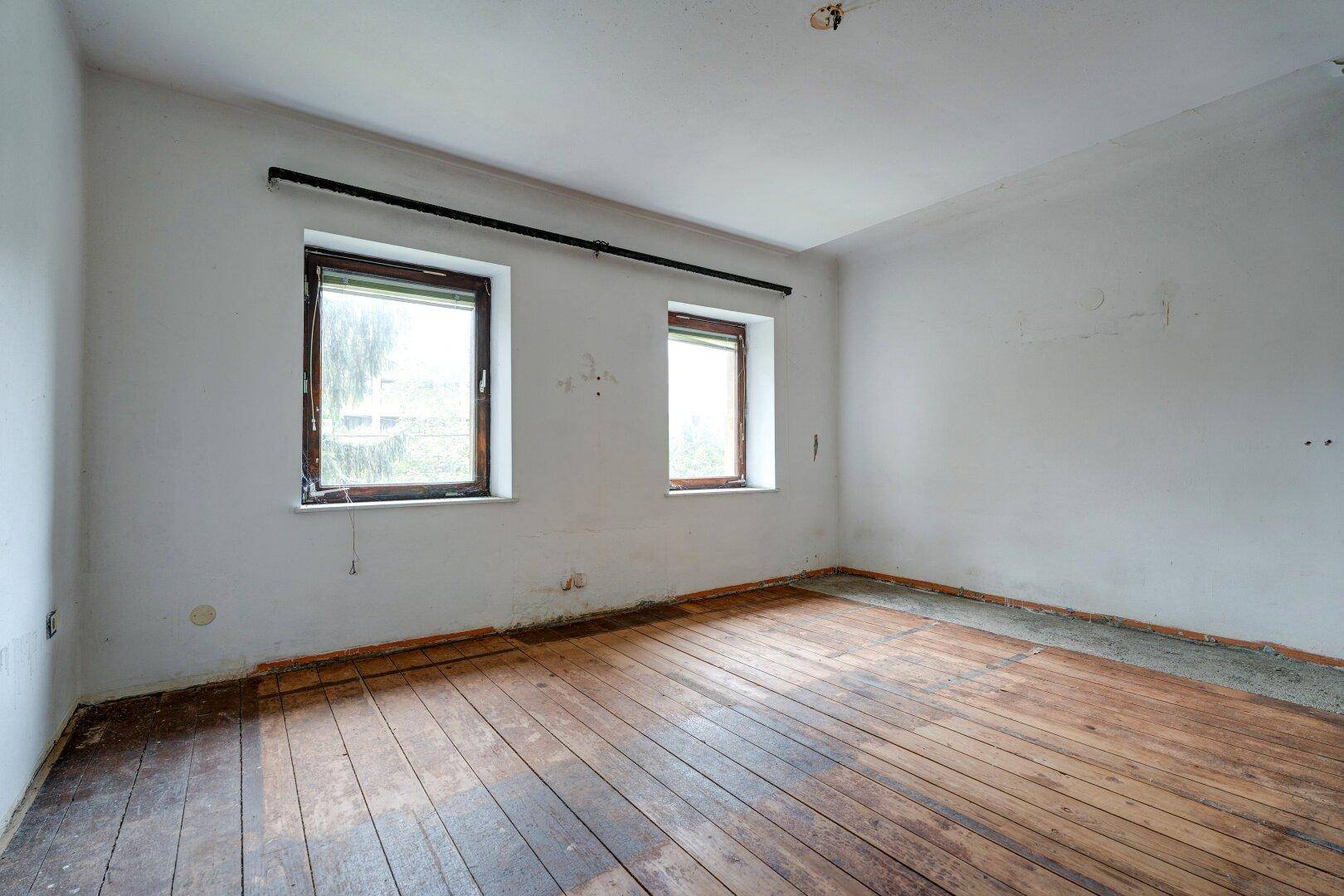 Leeres Zimmer mit Holzdielenboden, weißen Wänden und zwei Fenstern.