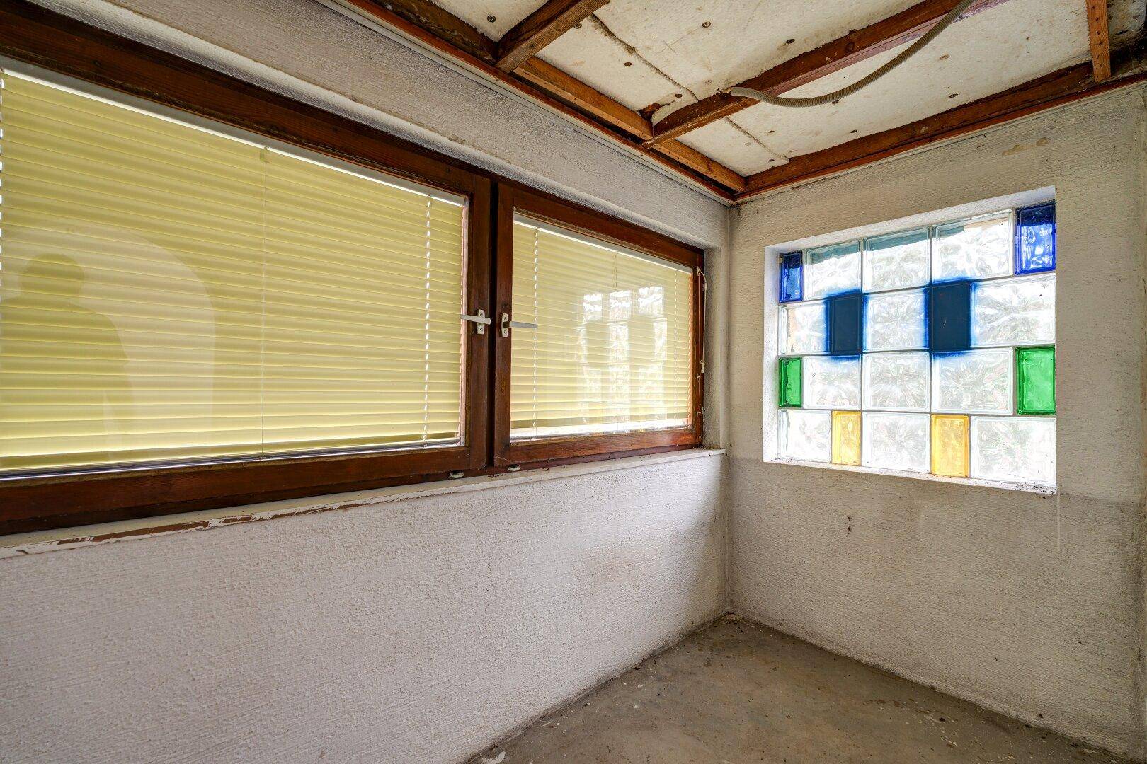 Kleiner Raum mit Buntglasfenster, Jalousien und sichtbarer Deckenkonstruktion.