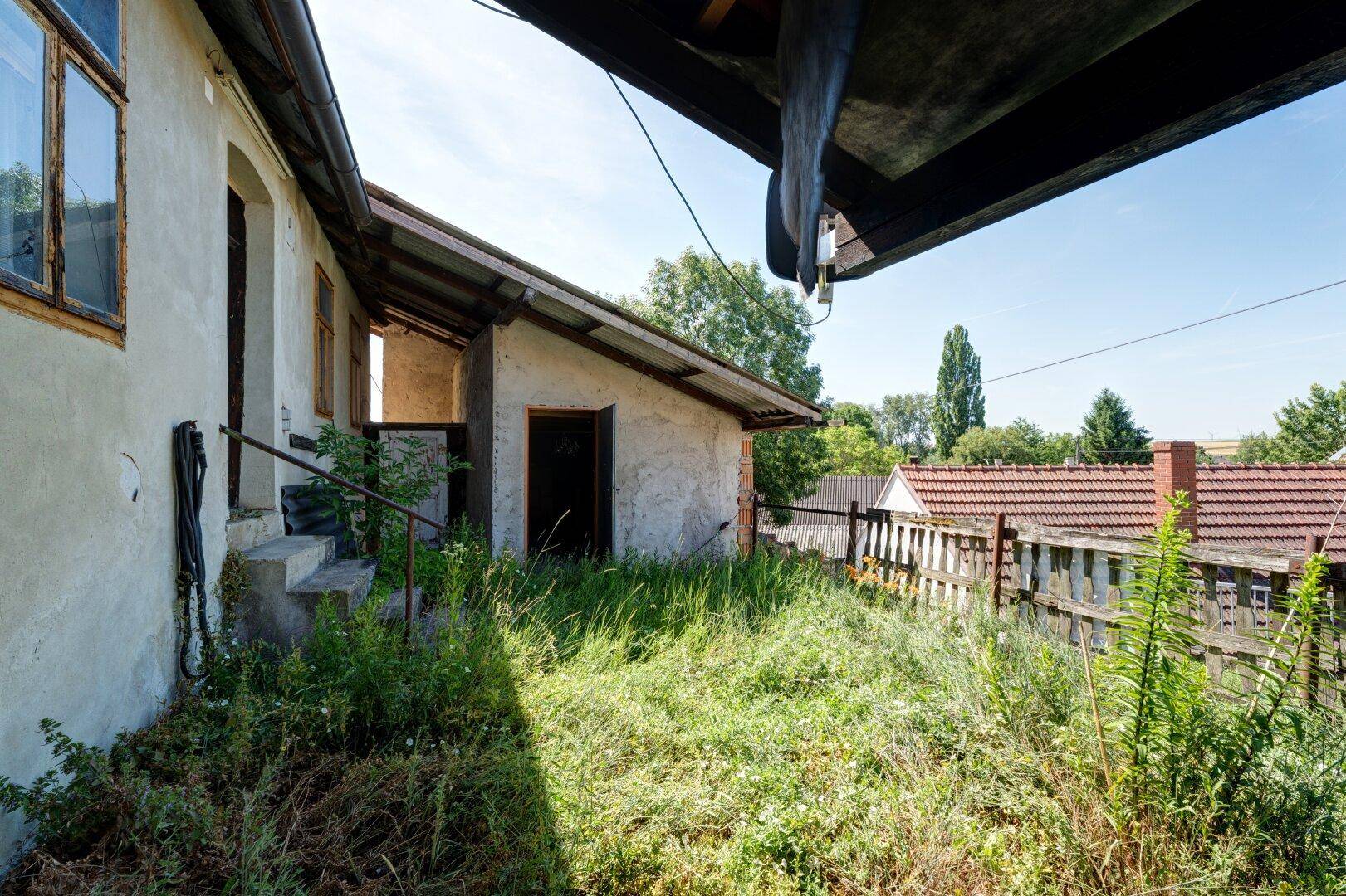 Hinterhof des Hauses mit hohem Gras, einem Holzzaun und Blick auf die ländliche Umgebung.