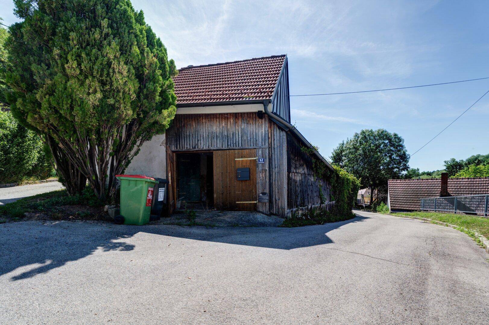 Scheunenartiges Gebäude mit großem Holztor, umgeben von Bäumen und einer kleinen Straße.