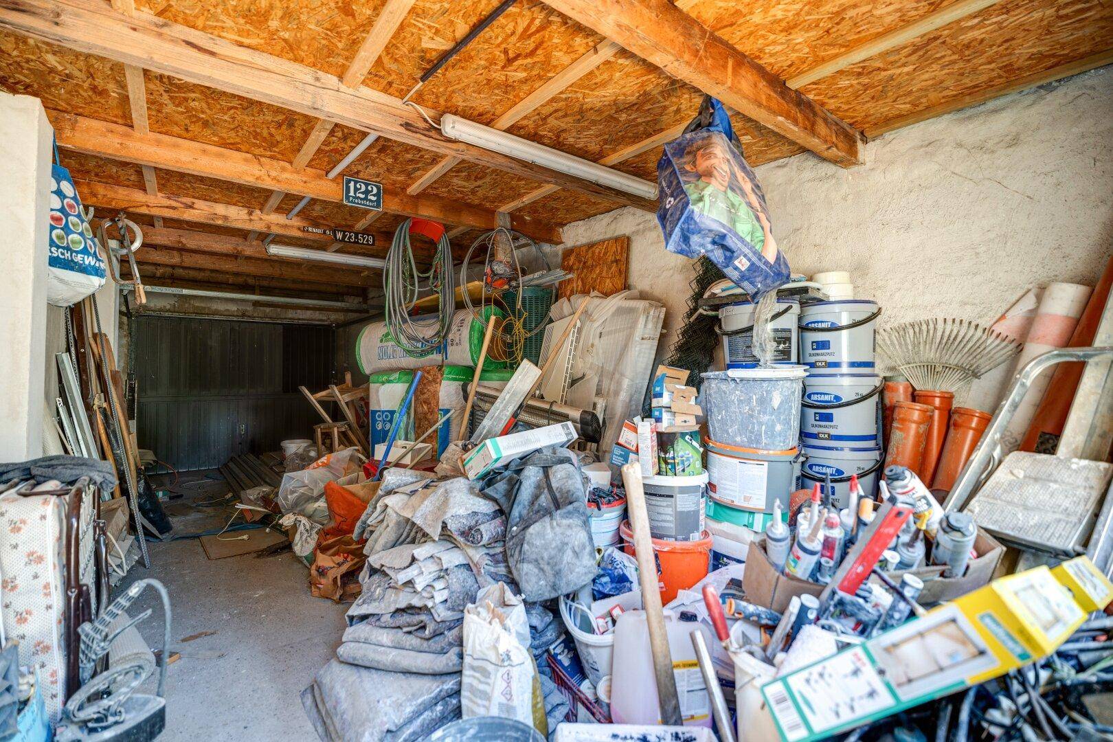 Unaufgeräumte Garage oder Abstellraum mit Werkzeugen, Farbeimern und sichtbaren Holzbalken.