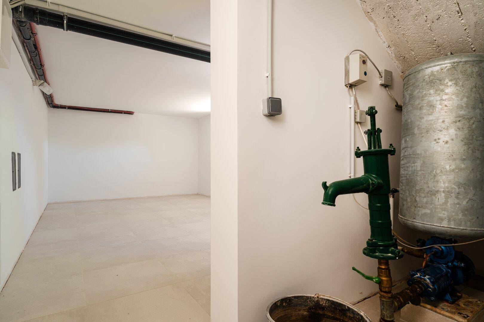 Kellerraum mit Fliesenboden, sichtbaren Leitungen und einer Wasserpumpe.