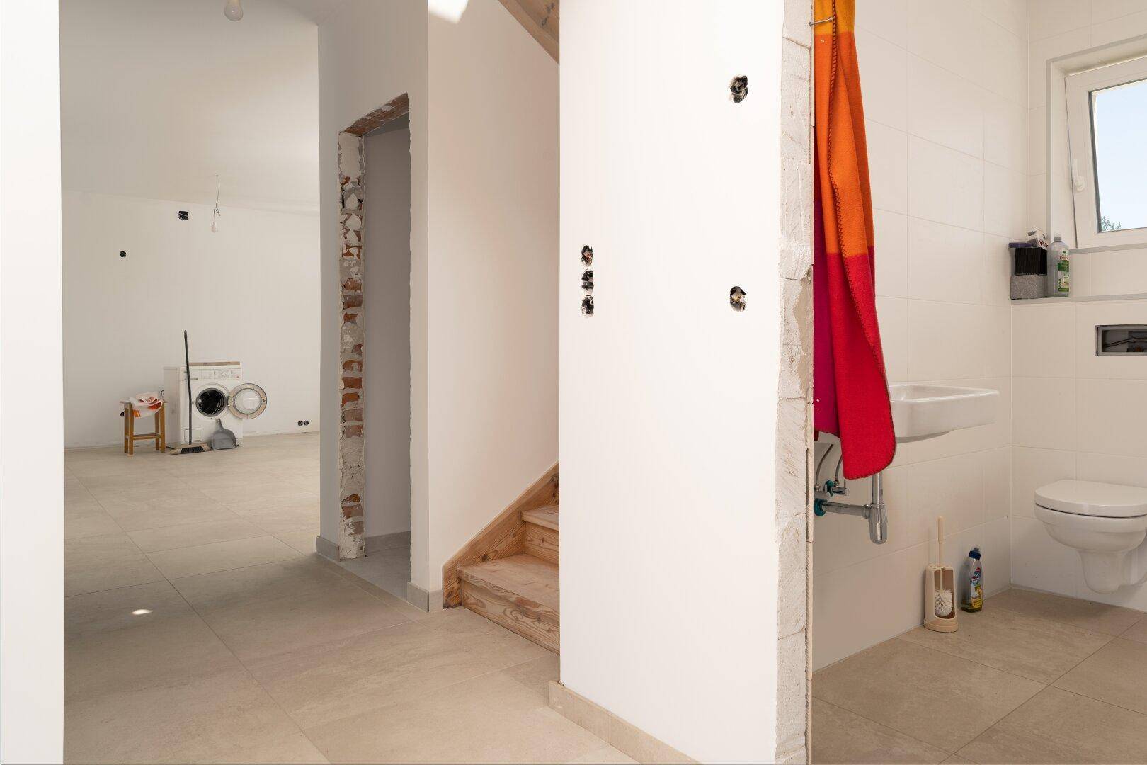 Flur mit Holztreppe und Blick auf ein Badezimmer sowie einen Raum im Rohbauzustand.