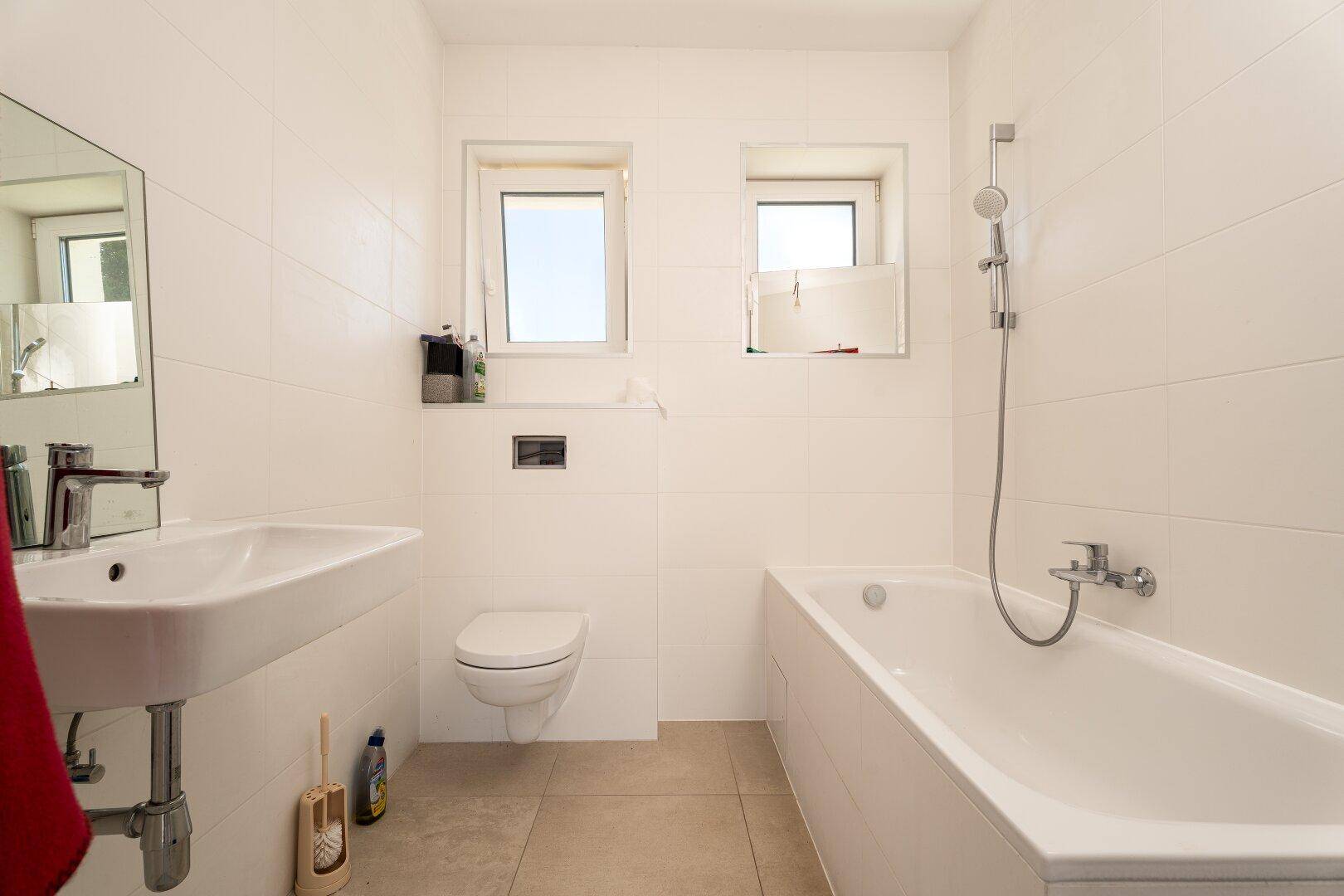 Modernes Badezimmer mit weißen Fliesen, Badewanne, WC, Waschbecken und zwei Fenstern.