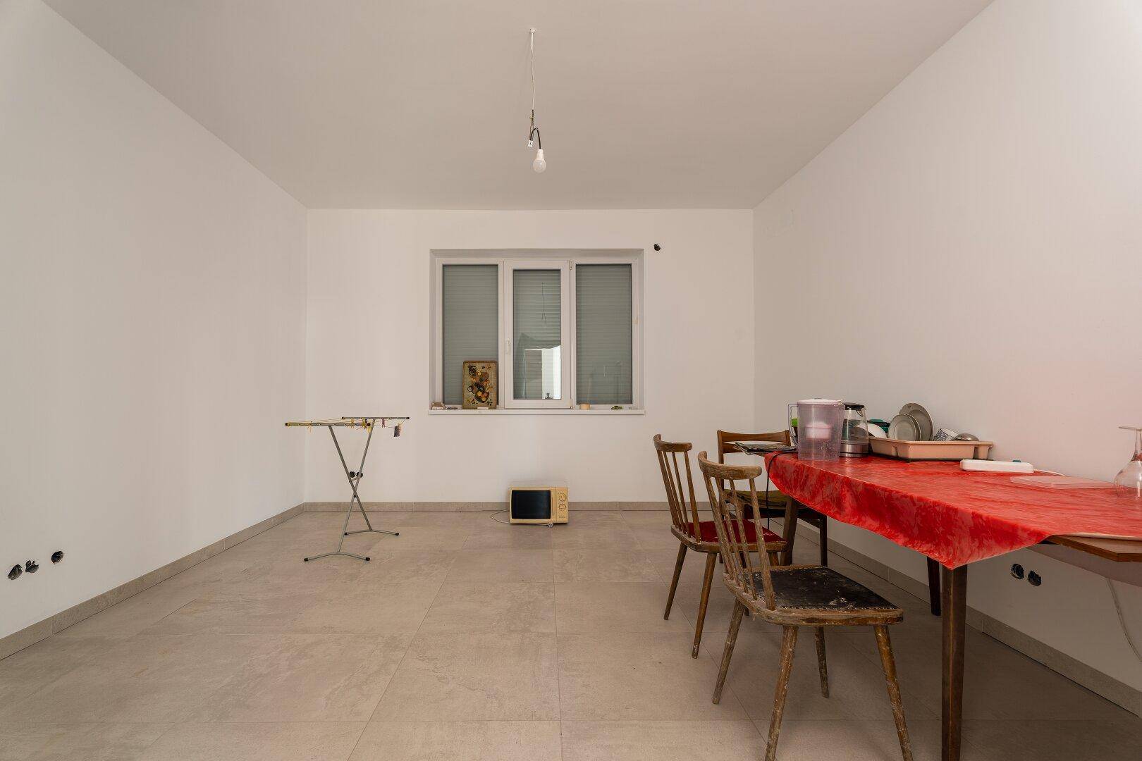 Geräumiger Raum mit Fliesenboden, Fenster und provisorischer Einrichtung, möglicherweise ein Esszimmer.