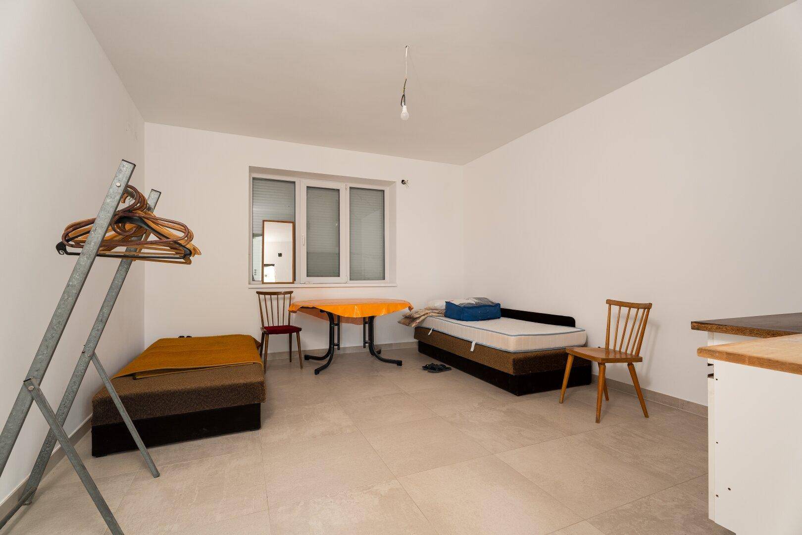 Helles Schlafzimmer mit Fliesenboden, Fenster und einfacher, provisorischer Möblierung.