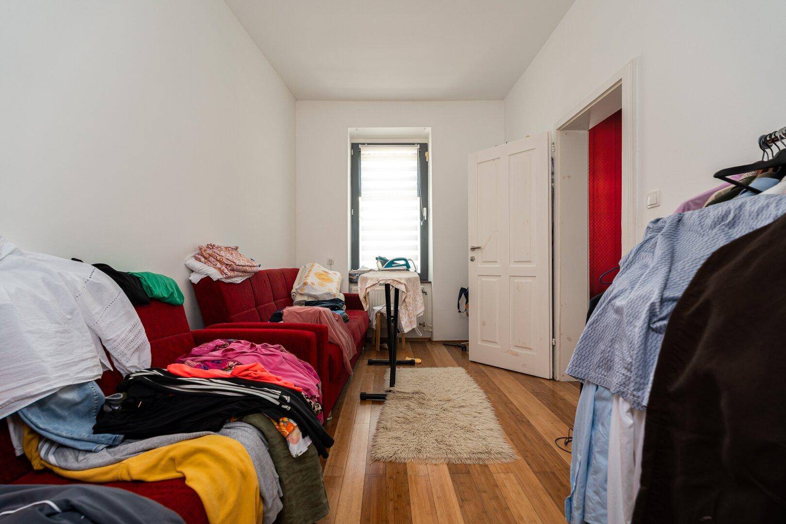 Kleiner Raum mit Sofa, Fenster und Kleiderstange, ideal als Gäste- oder Abstellraum.
