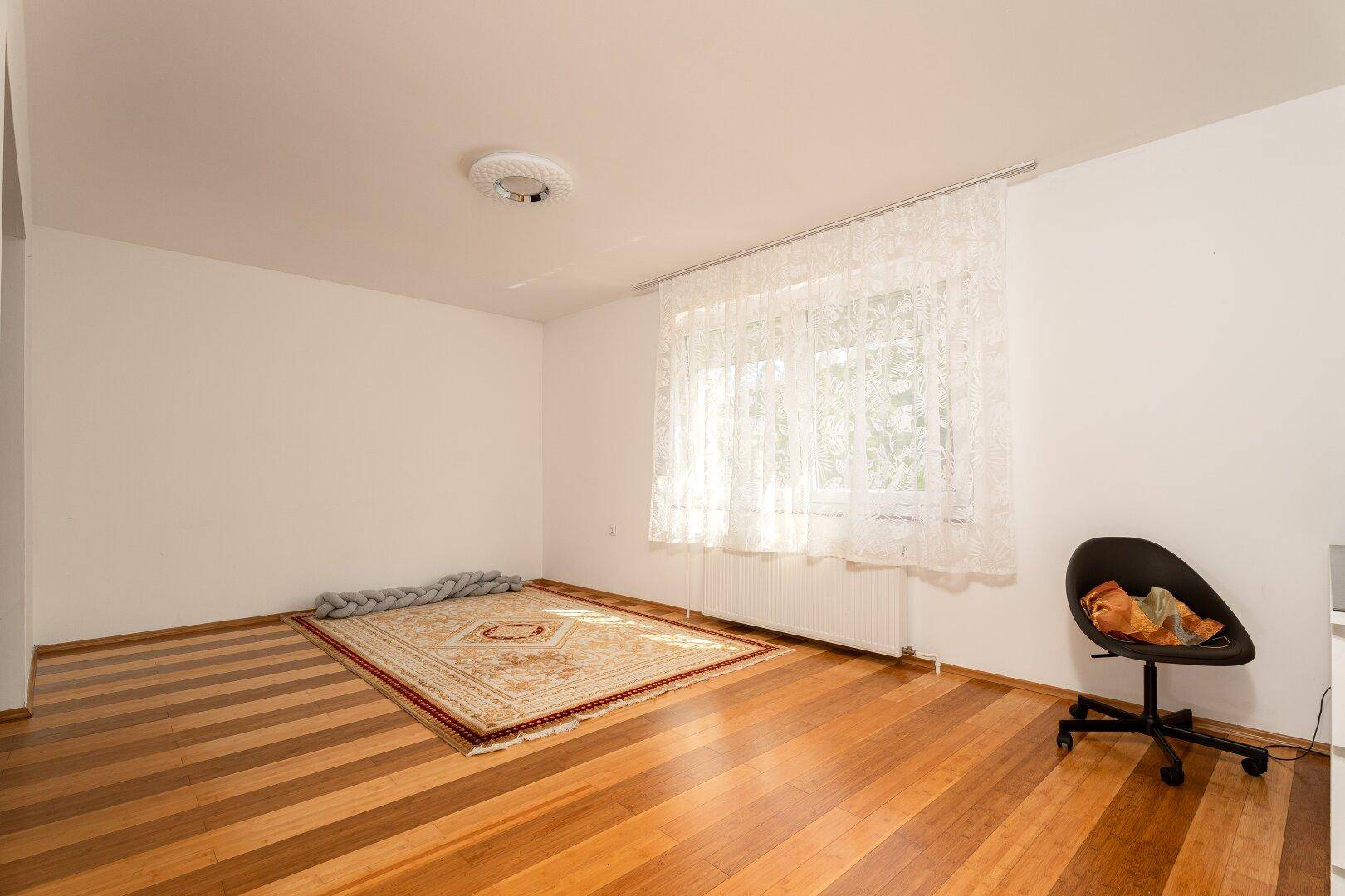 Helles Zimmer mit breiten Holzplankenböden, weißen Wänden und einem Fenster mit Vorhängen.