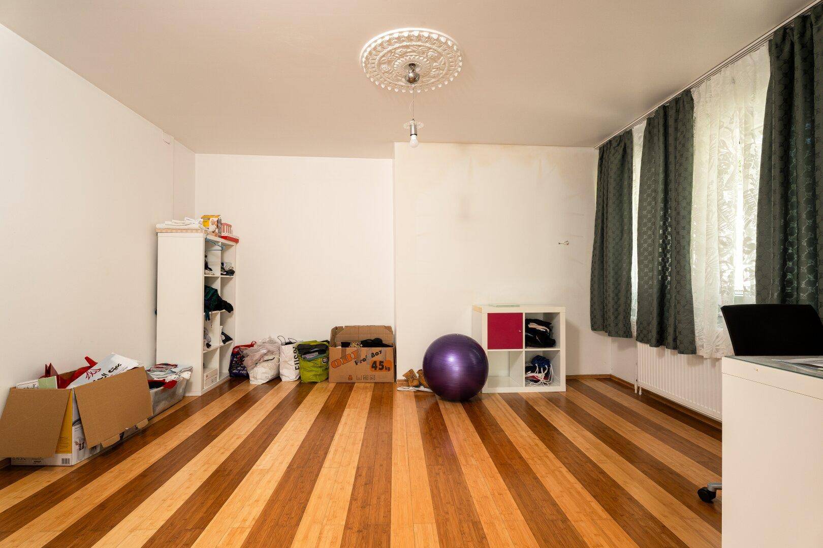 Geräumiges Zimmer mit Holzplankenboden, Stuckdecke und einem Fenster mit dunklen Vorhängen.