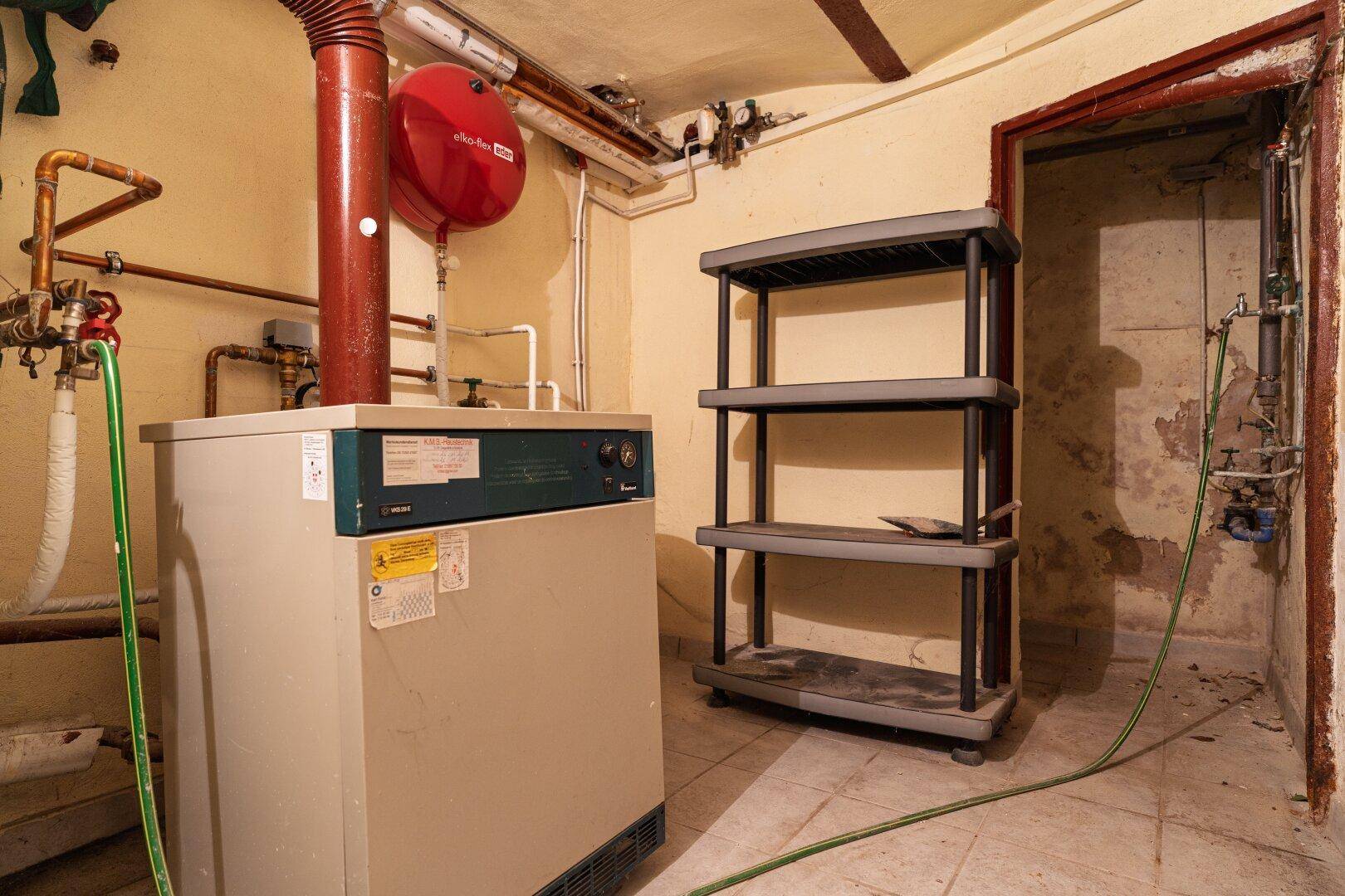 Heizungsraum im Keller mit Boiler, Rohren und einem Regal, zeigt den technischen Bereich des Hauses.