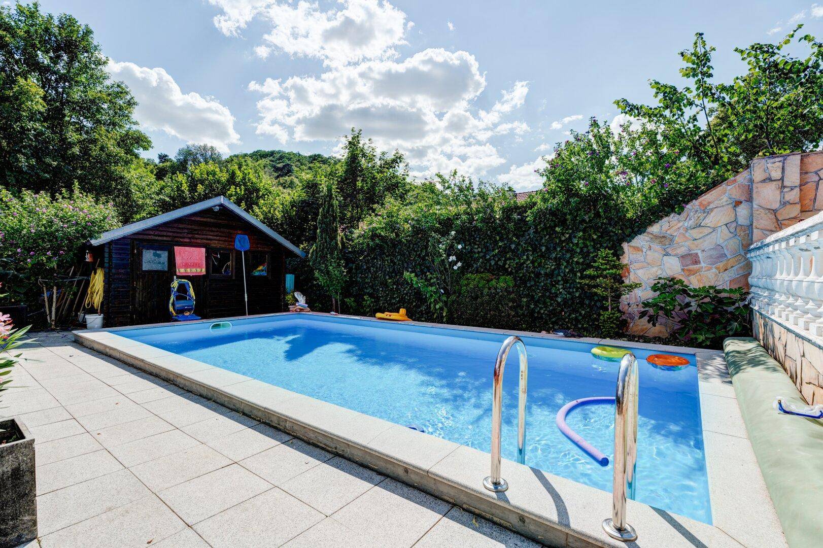 Blauer Swimmingpool im Gartenbereich mit Holzschuppen und Natursteinmauer.