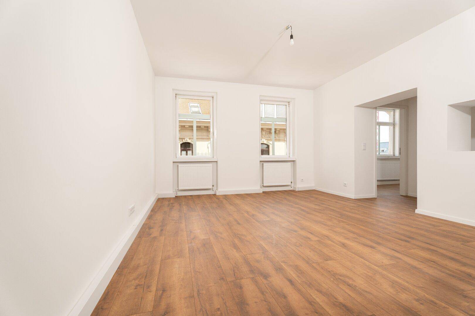 Geräumiges Wohnzimmer mit hellem Laminatboden und zwei großen Fenstern.