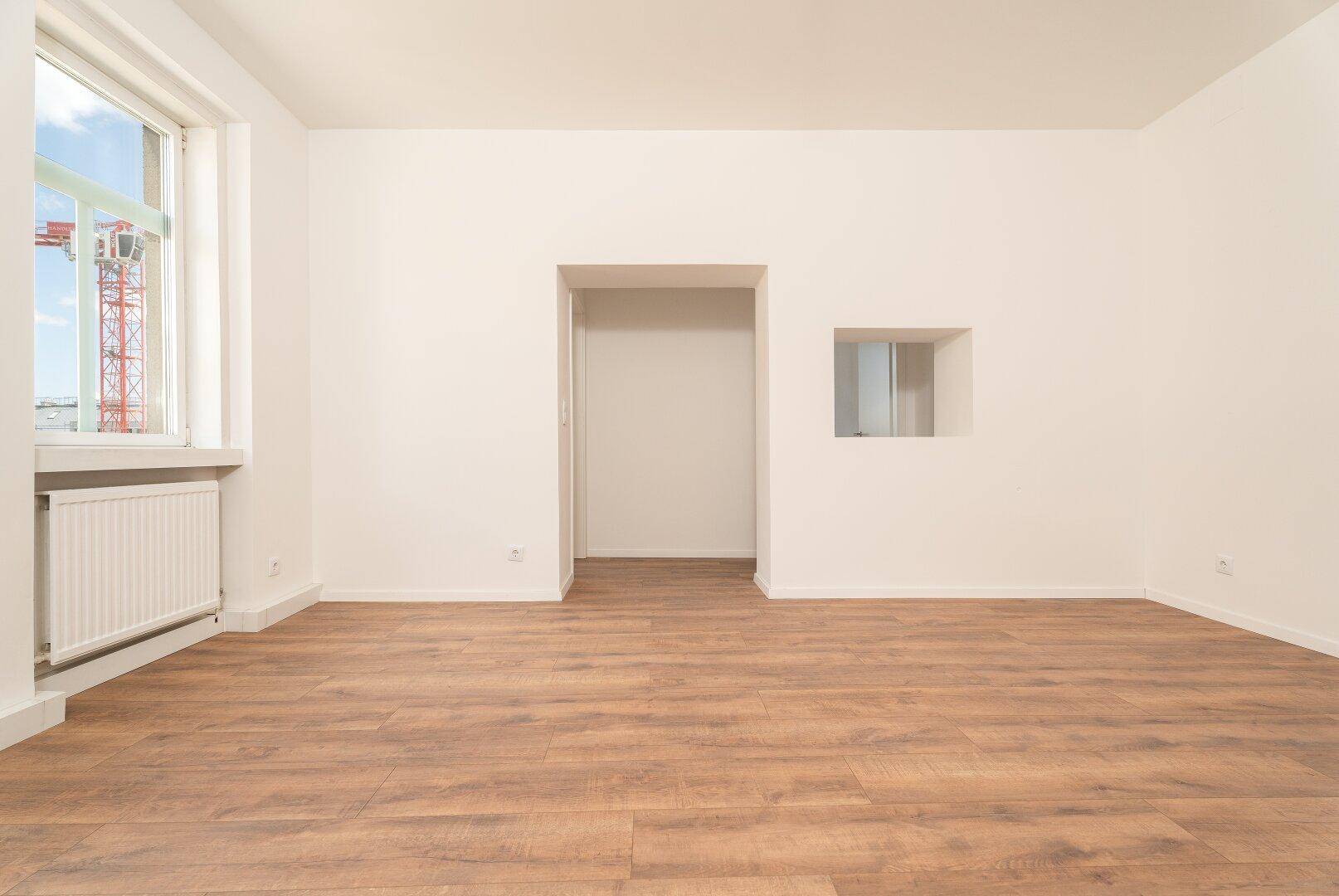 Heller Wohnbereich mit Laminatboden und einem Fenster, das Tageslicht hereinlässt.