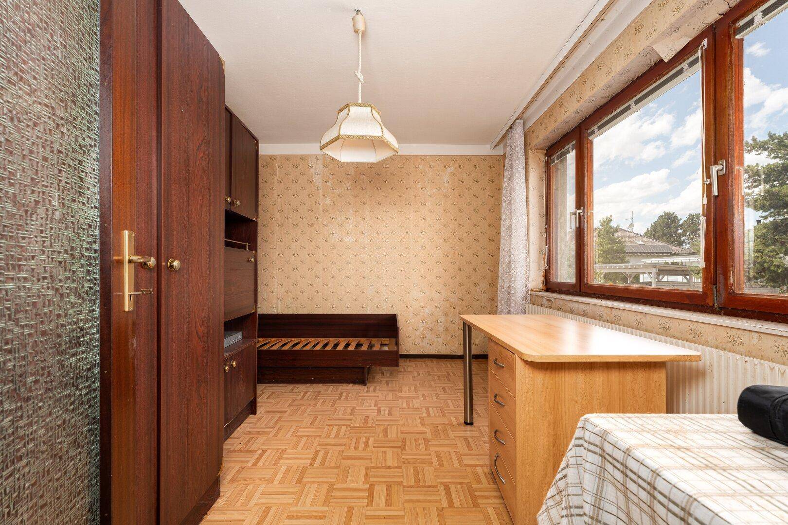Schlafzimmer oder Büro mit Holzmöbeln, gemusterter Tapete und Fenster.