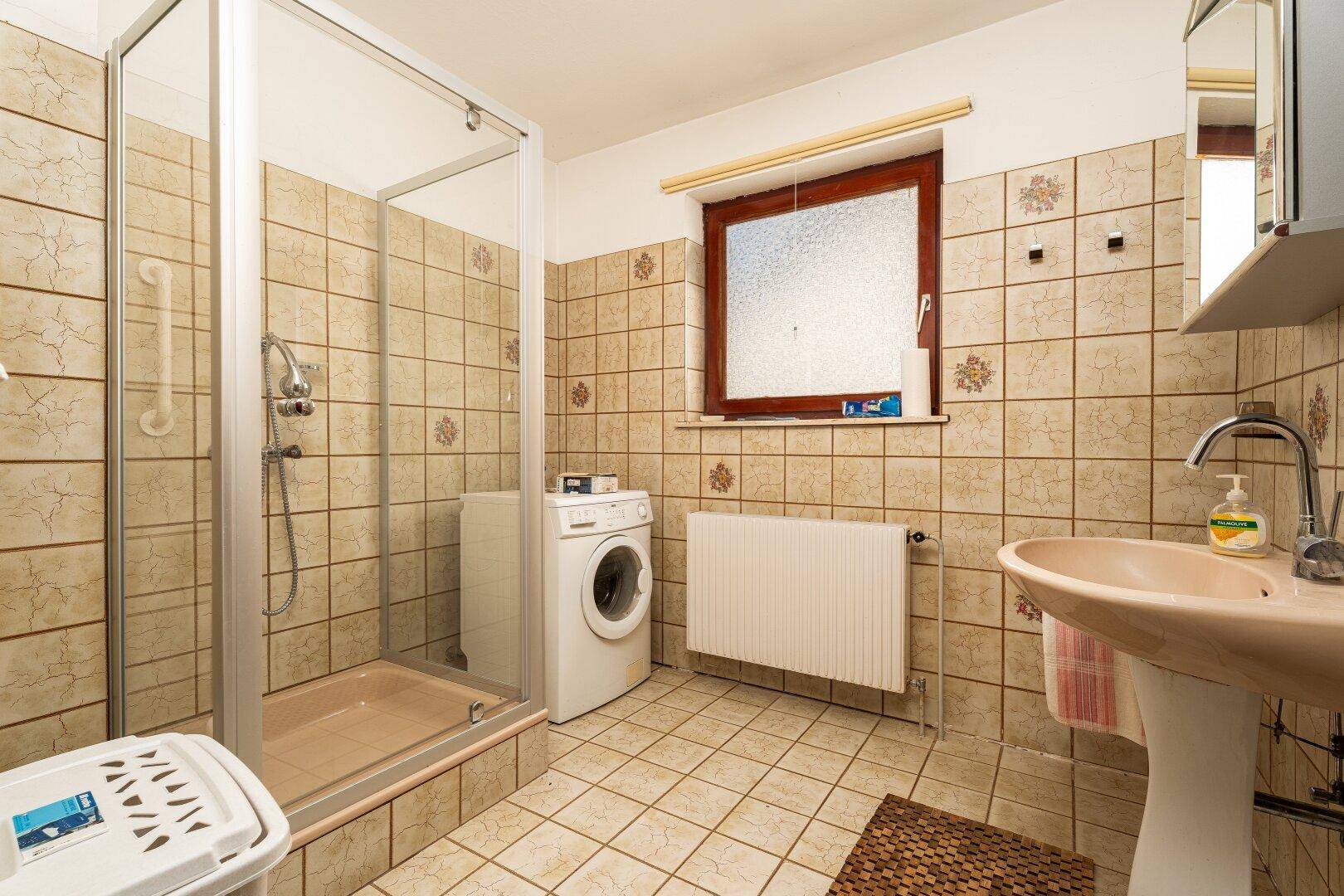 Badezimmer mit Dusche, Waschbecken, Waschmaschine und beigefarbenen Fliesen.