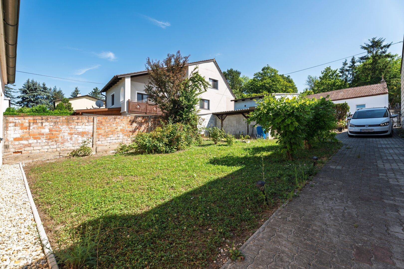 Sonniger Gartenbereich mit Rasenfläche, alter Ziegelmauer und Blick auf das Nachbarhaus.