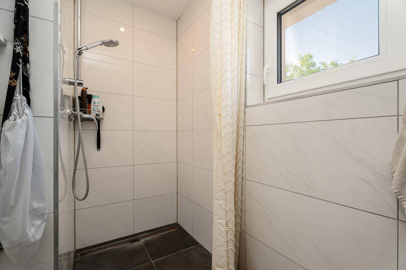 Duschbereich im Badezimmer mit weißen Fliesen, Duschvorhang und Fenster für Tageslicht.