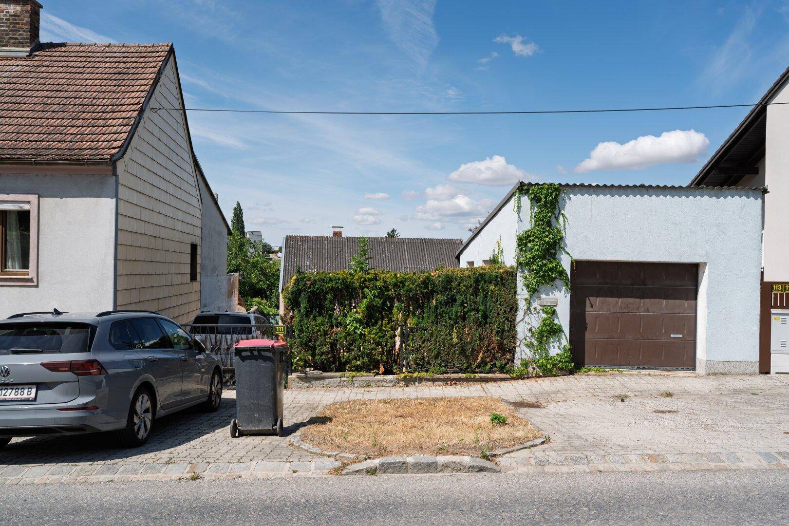 Ansicht von der Straße mit geparktem Auto, einer Garage und Hecke unter blauem Himmel.