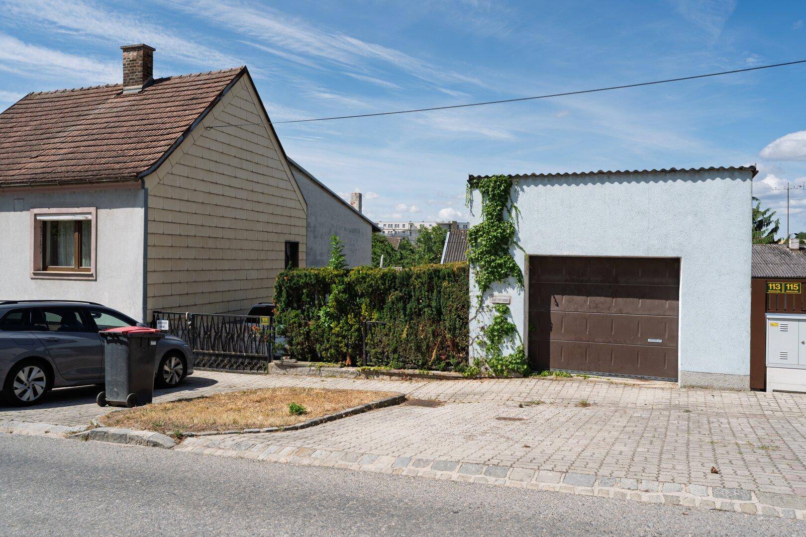 Ansicht der Garage mit braunem Tor und dem angrenzenden Wohnhaus von der Straße aus.