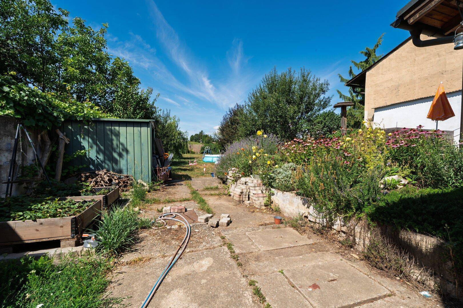 Länglicher Gartenweg mit Hochbeeten, Gartenhaus und einem kleinen Aufstellpool im Hintergrund.