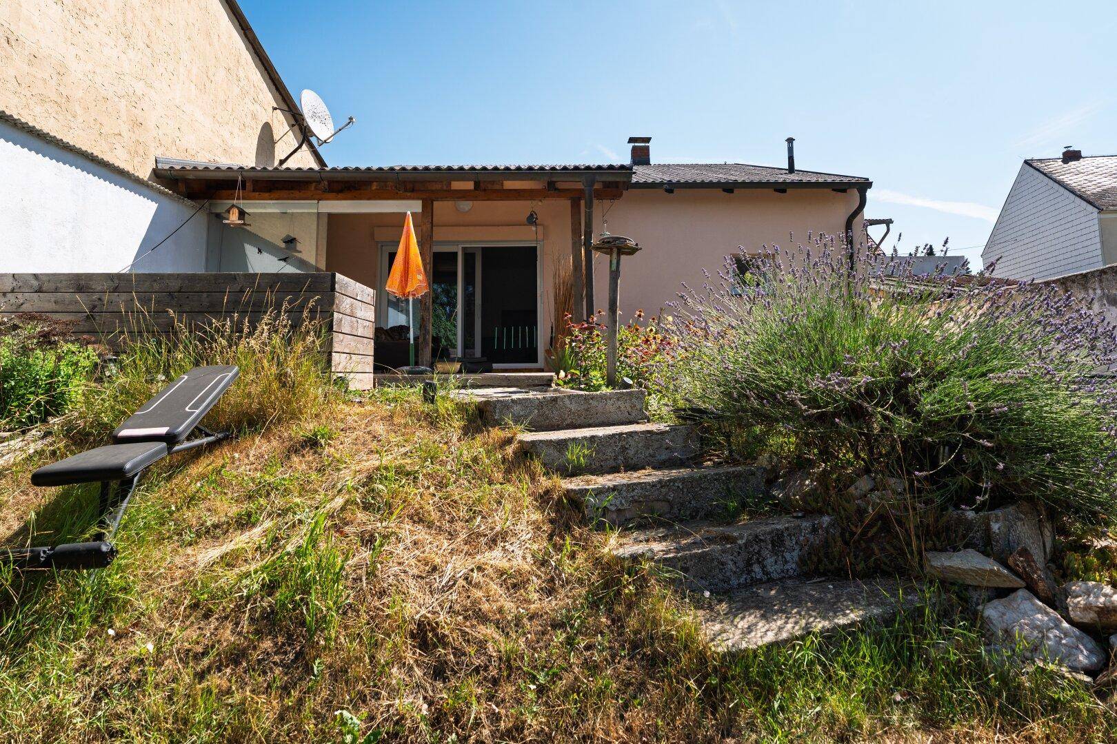 Überdachte Terrasse am Haus mit Außentreppe, umgeben von einem naturbelassenen Gartenbereich.