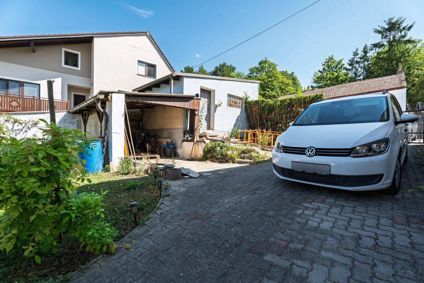 Außenansicht des Hauses mit gepflasterter Einfahrt, weißem Auto und einem überdachten Unterstand.