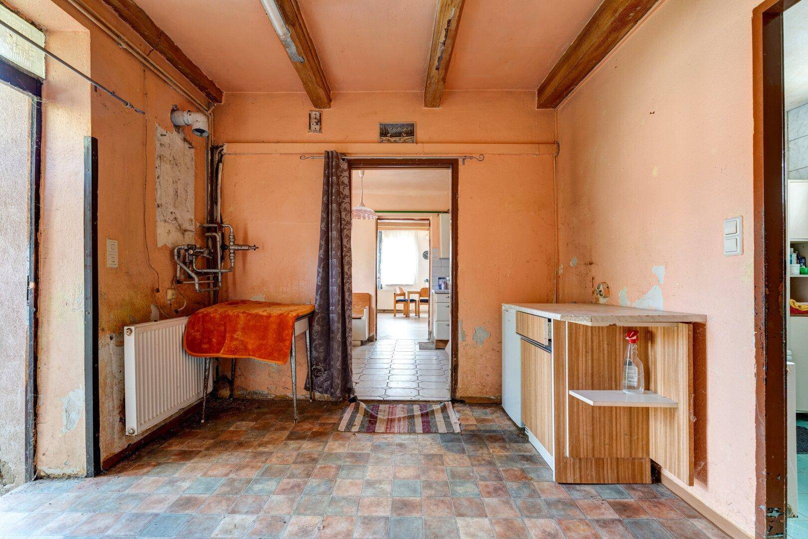 Einfacher Raum mit sichtbaren Holzbalken, Fliesenboden und Heizkörper, möglicherweise ein Hauswirtschaftsraum.
