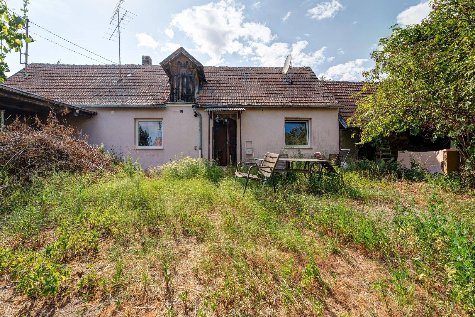 Rückseite des Hauses mit einem verwilderten Garten und einer alten Sitzgruppe im Freien.