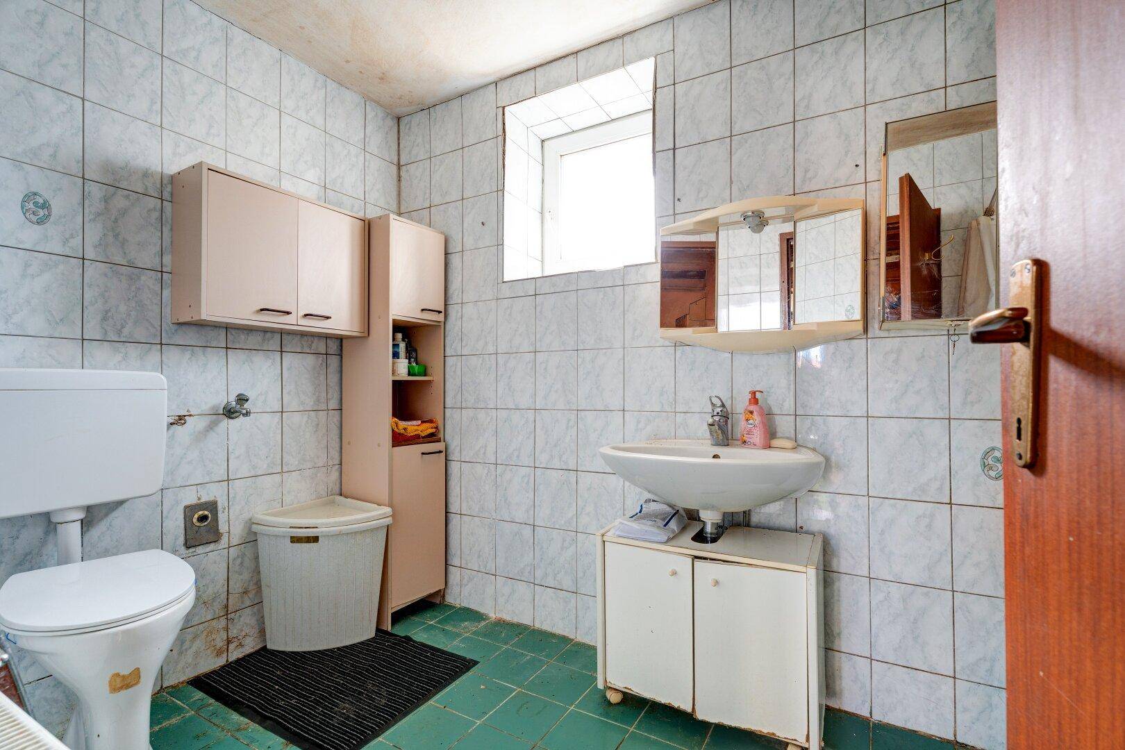 Badezimmer im Stil der 80er Jahre mit weißen Fliesen, Waschbecken, Toilette und kleinem Fenster.