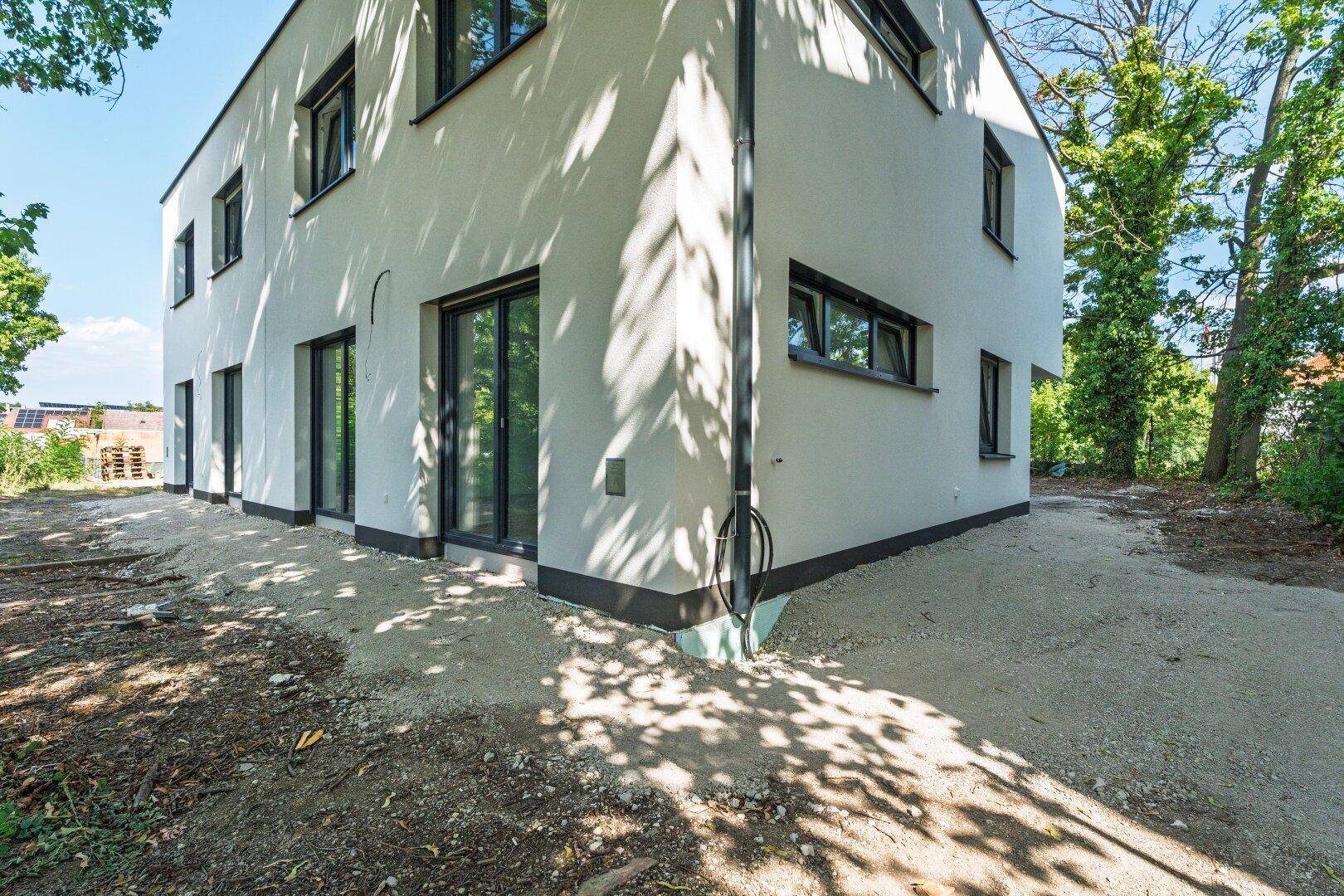 Seitenansicht eines modernen Neubaus mit weißer Fassade und dunklen Fensterrahmen, umgeben von Bäumen.
