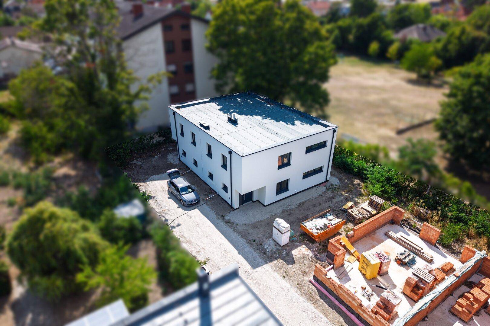 Luftaufnahme eines modernen Hauses mit Flachdach und angrenzender Baustelle für weitere Gebäude in ländlicher Umgebung.