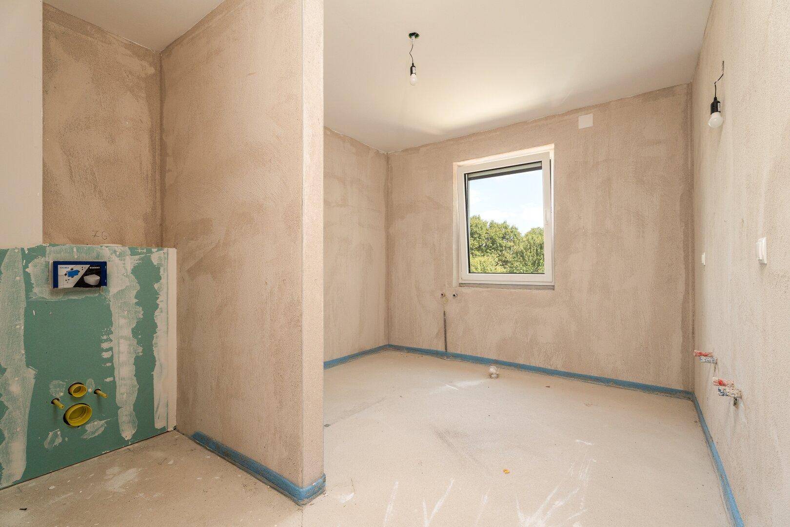 Badezimmer im Rohbau mit Fenster und vorbereiteten Anschlüssen für Sanitärinstallationen, bereit für den Ausbau.