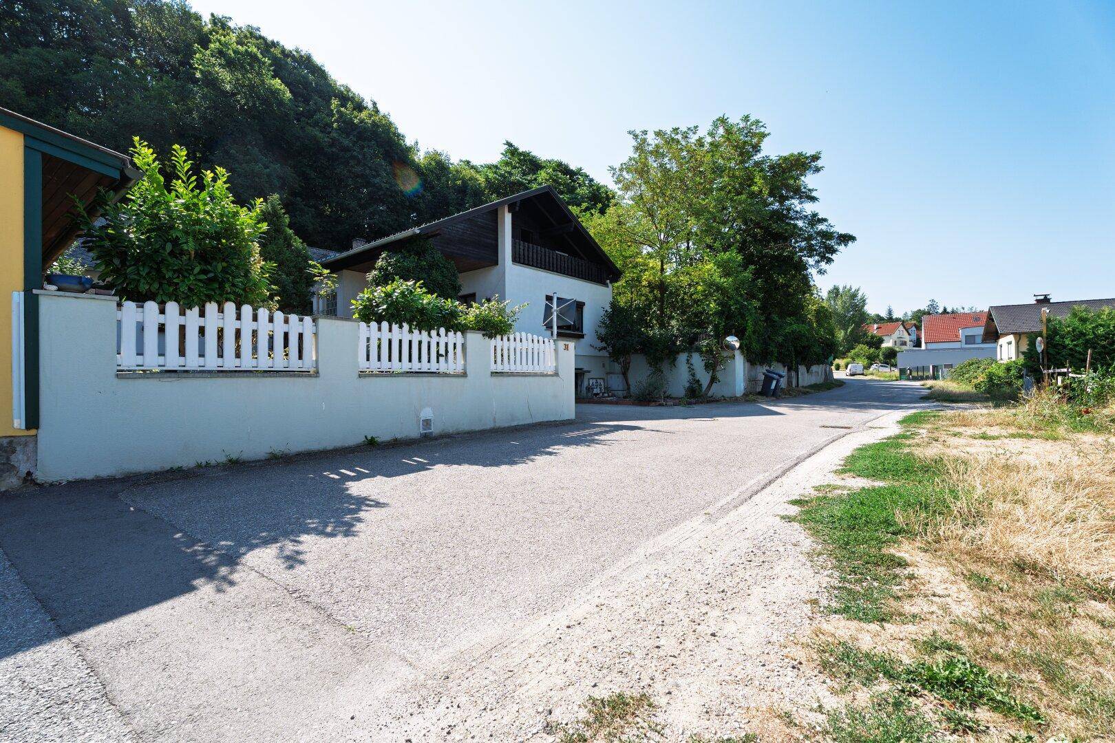 Hausansicht von der Straße mit weißem Zaun und umgebender Natur in ländlicher Umgebung.
