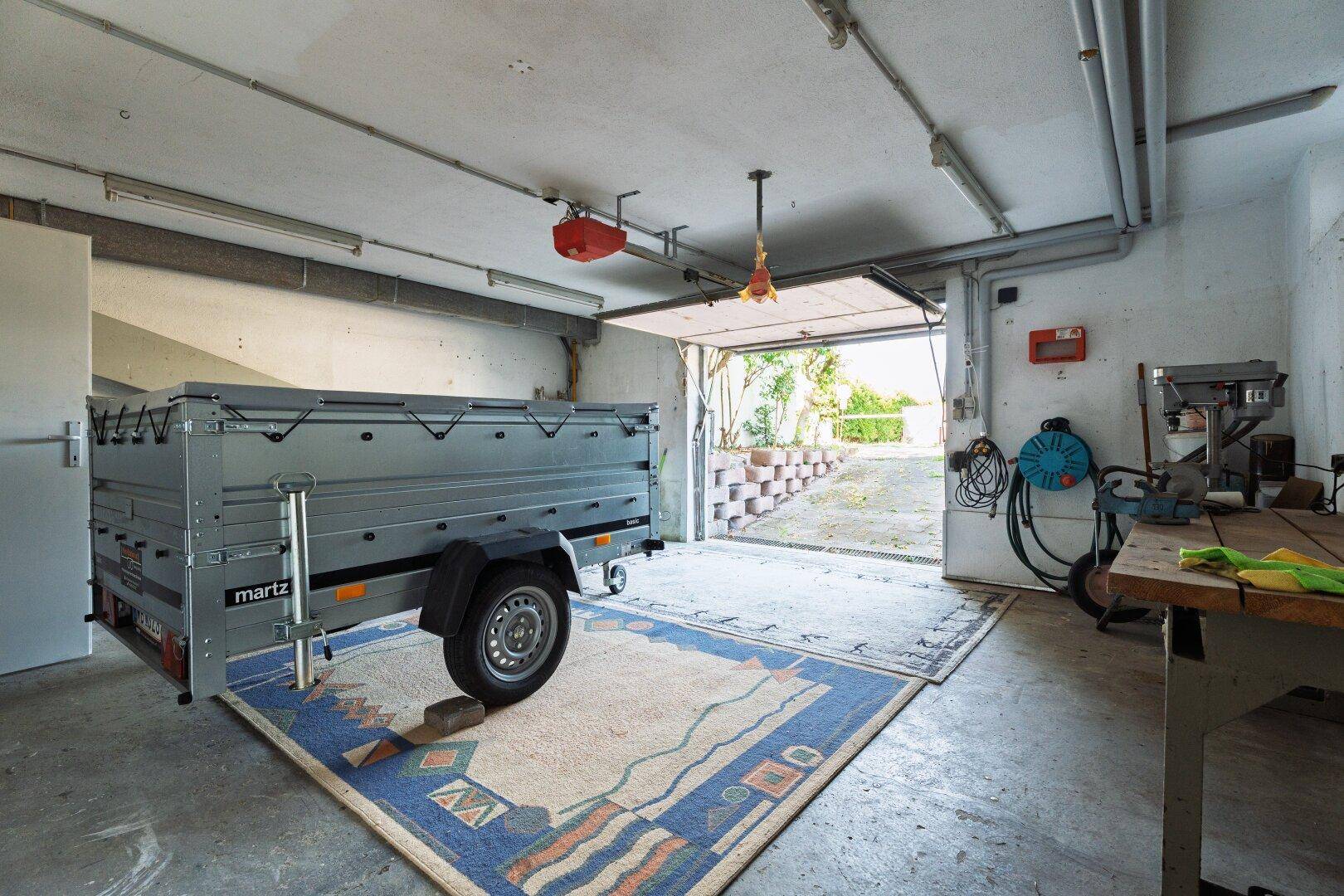 Geräumige Garage mit offenem Tor, einem Anhänger und einer Werkbank, ideal für Fahrzeuge und Hobby.