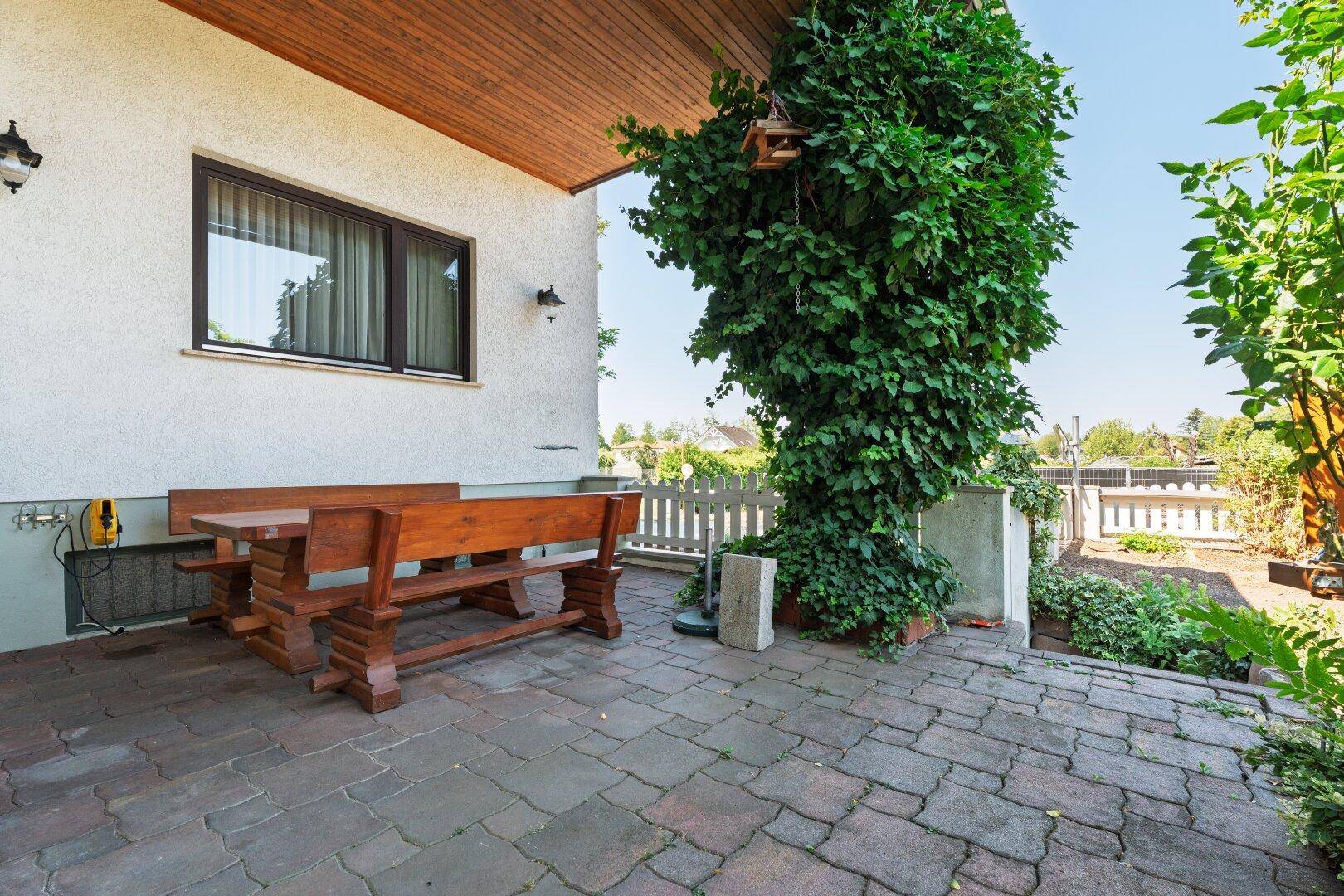 Gemütliche Terrasse mit Holztisch und Sitzbänken, umgeben von üppigem Grün und Kletterpflanzen.