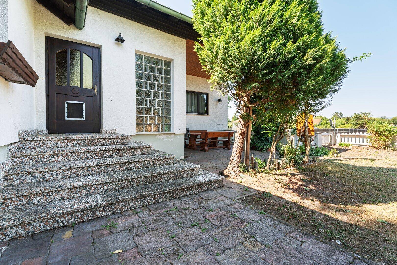 Eingangsbereich mit Steintreppe und gepflastertem Vorplatz, daneben ein kleiner Gartenbereich.