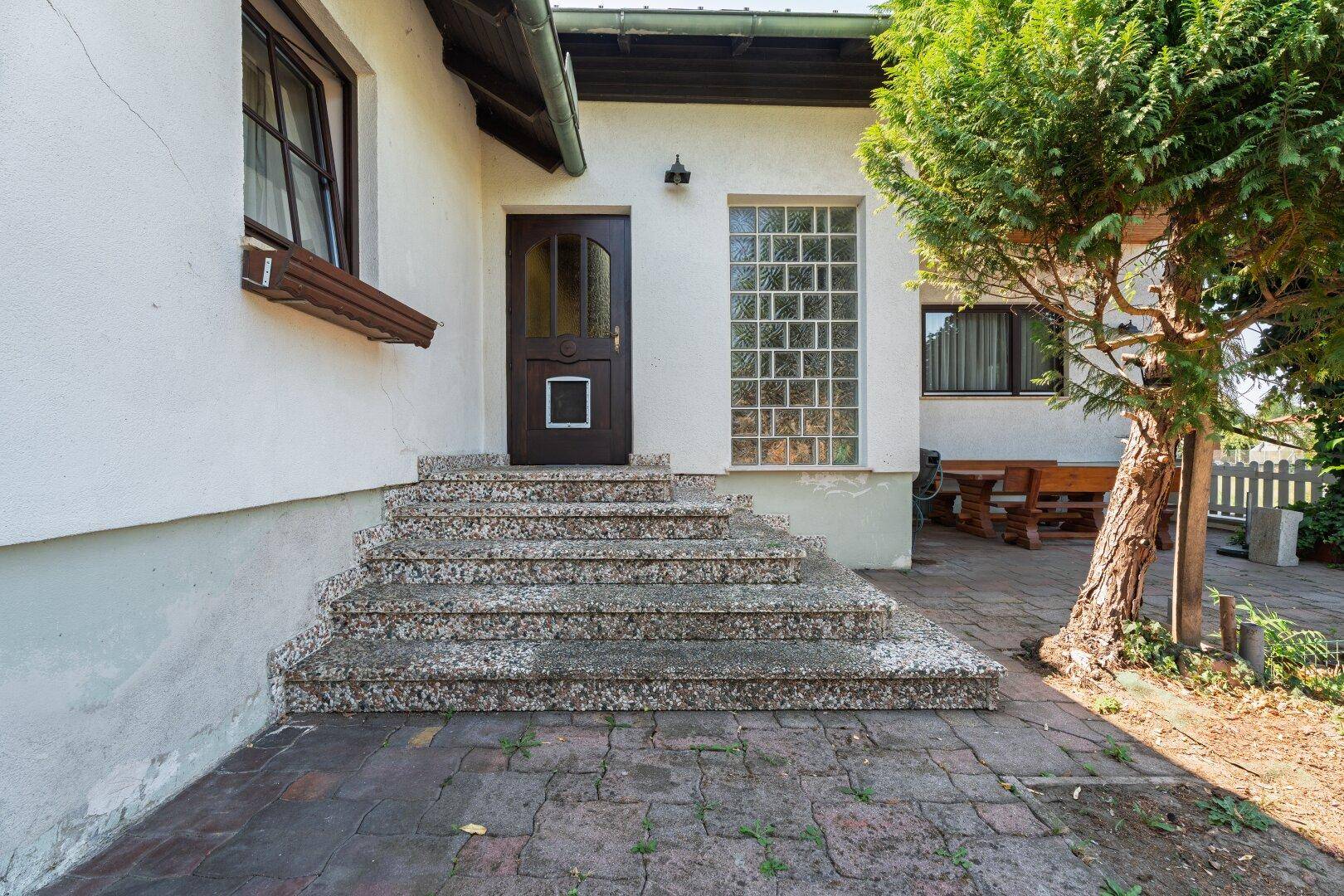 Eingangsbereich des Hauses mit Steintreppe und Holztür, umgeben von einem kleinen Baum.