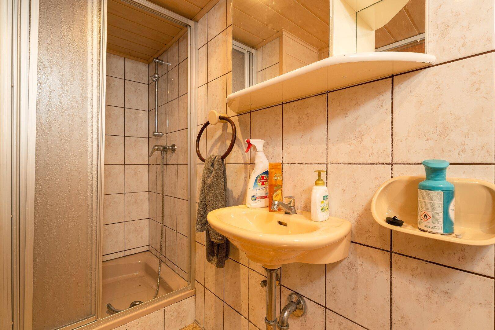 Kleines Badezimmer mit Dusche, Waschbecken und beigefarbenen Fliesen im 80er-Jahre-Stil.
