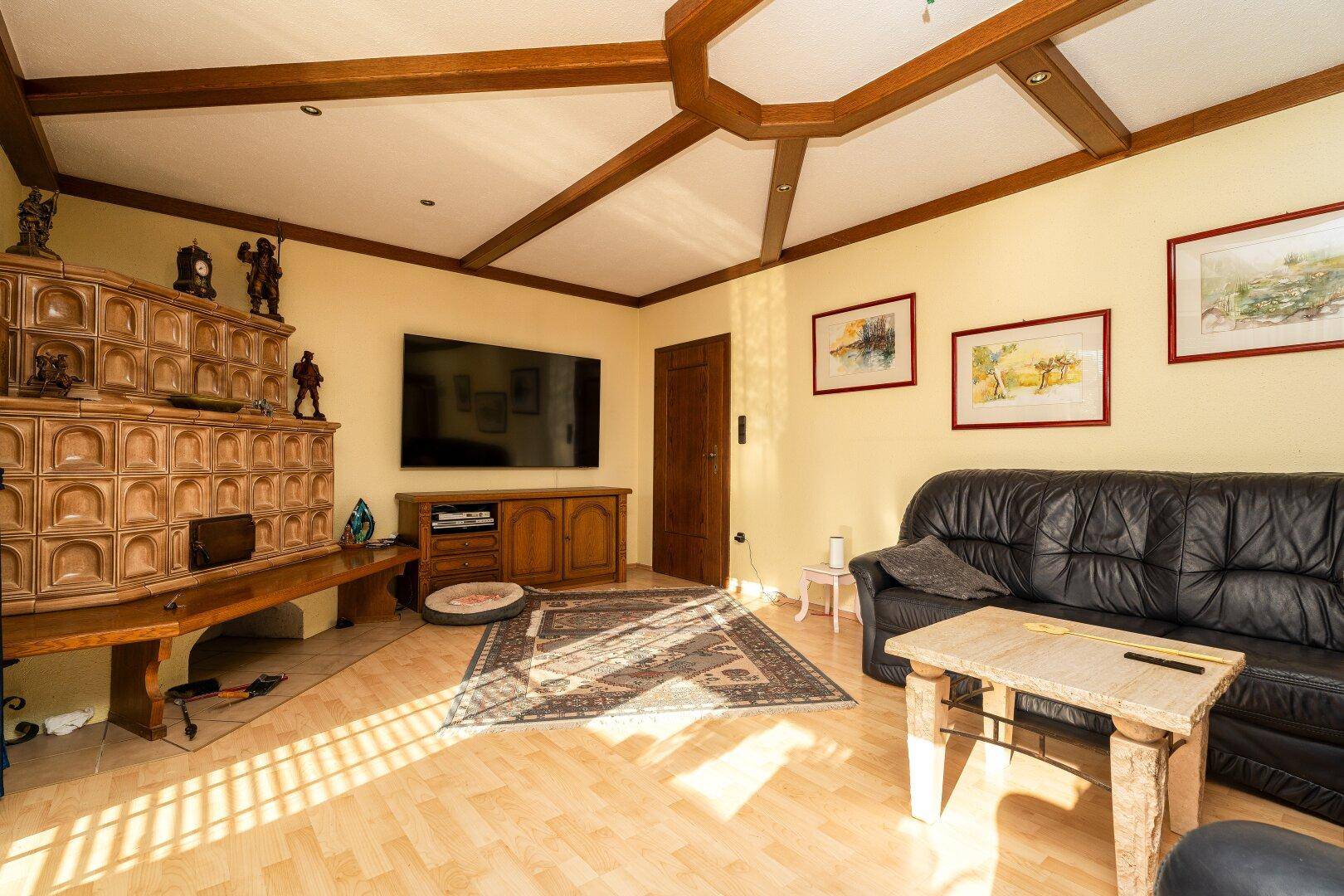 Geräumiges Wohnzimmer mit Kachelofen, Holzbalkendecke und Parkettboden in hellem Ambiente.