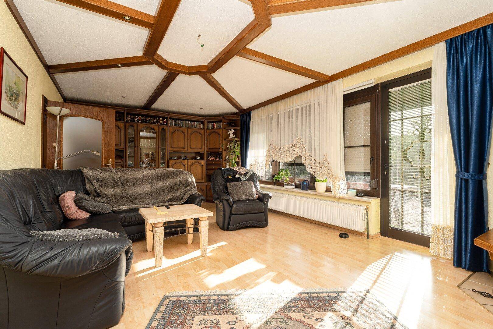 Helles Wohnzimmer mit Ledersofa, Holzbalkendecke und Zugang zur Terrasse durch große Fenster.