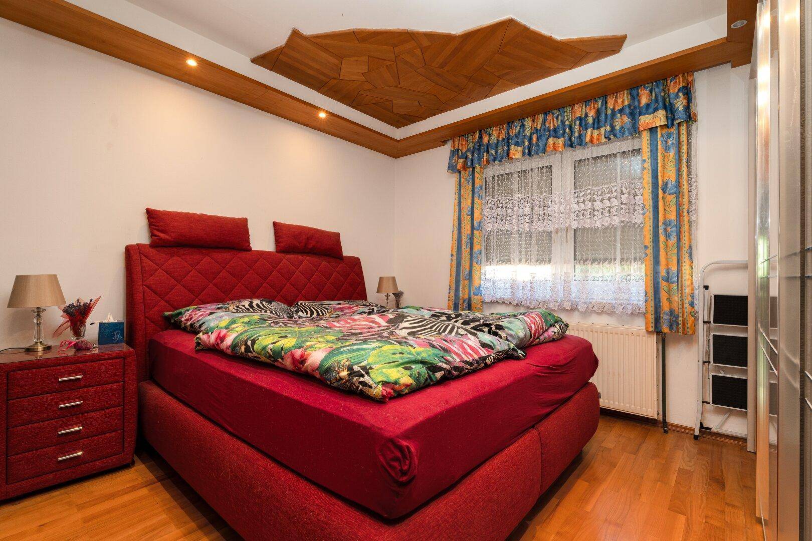 Gemütliches Schlafzimmer mit rotem Bett, Holzboden und dekorativer Decke für eine warme Atmosphäre.