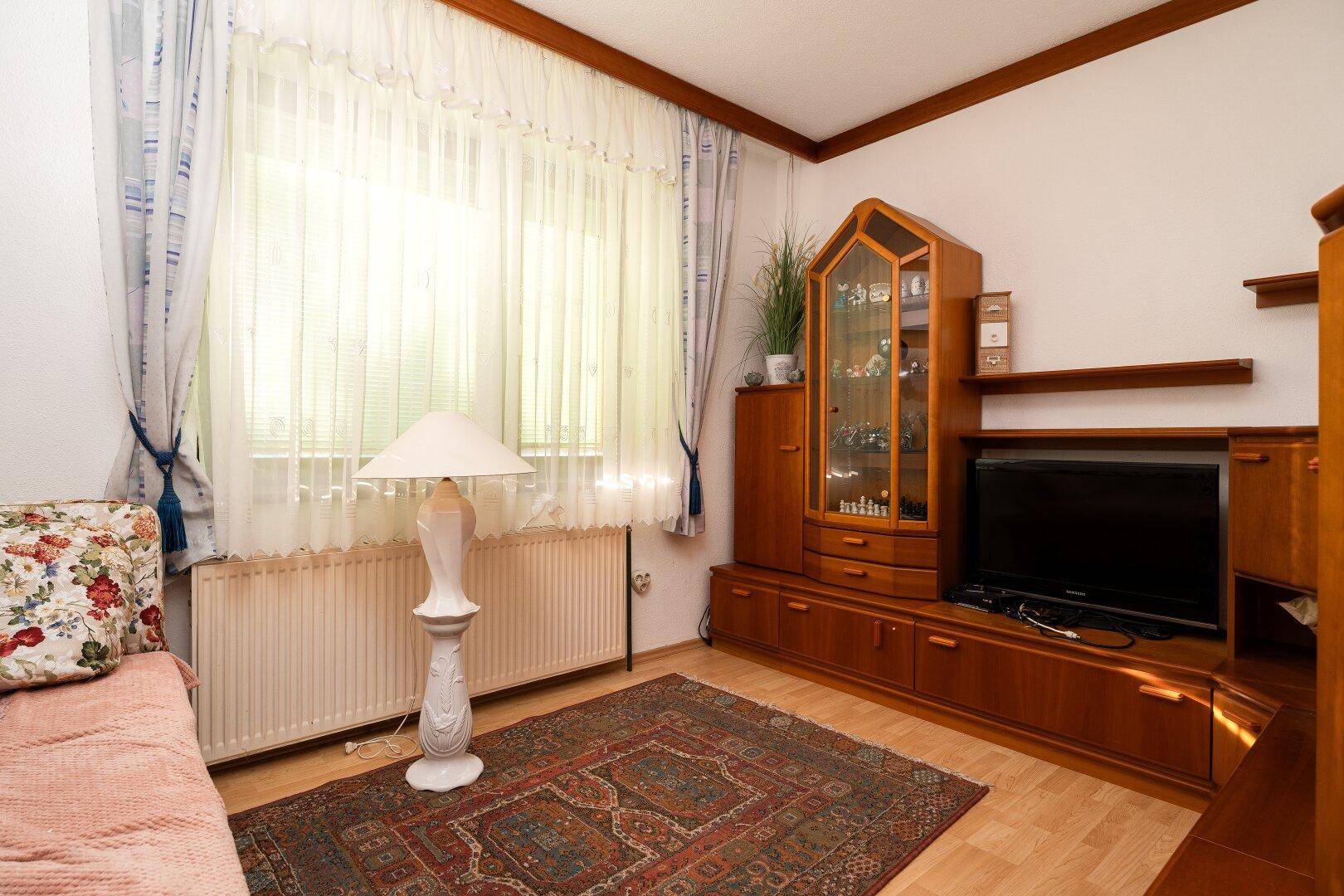 Helles Wohnzimmer mit Holzmöbeln, Teppich und großen Fenstern, die viel Licht hereinlassen.