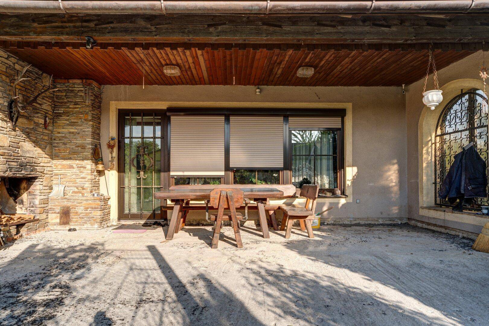 Überdachte Terrasse mit großem Holztisch, gemauertem Kamin und Rollläden an den Fenstern.