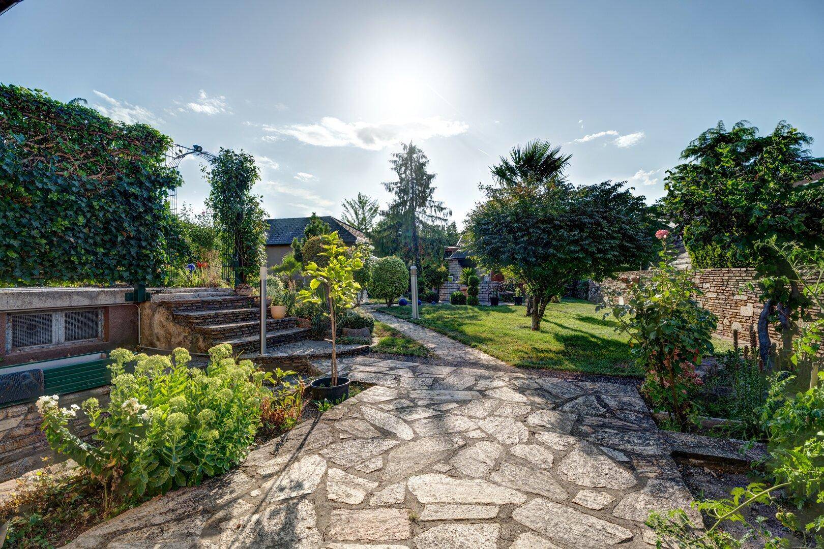 Weitläufiger Garten mit Steinwegen, Terrassen und vielfältiger Bepflanzung unter strahlender Sonne.