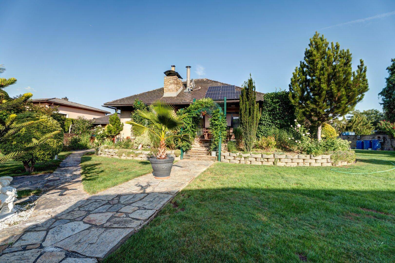 Weitläufiger Garten mit Steinwegen, Palmen und Blick auf das Haus unter blauem Himmel.